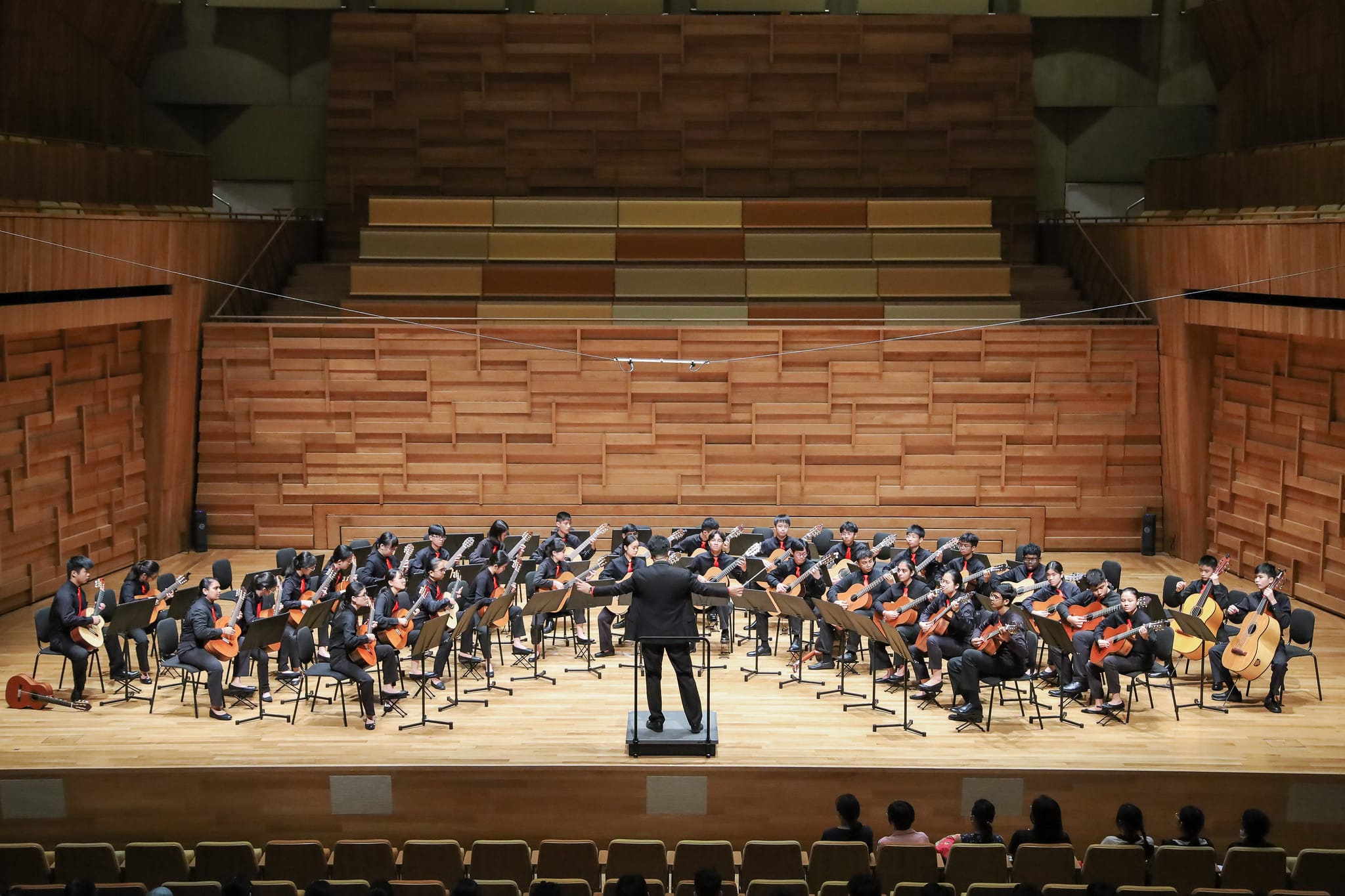 A conductor leads a large guitar ensemble in black uniforms with red ties on a wooden concert hall stage.